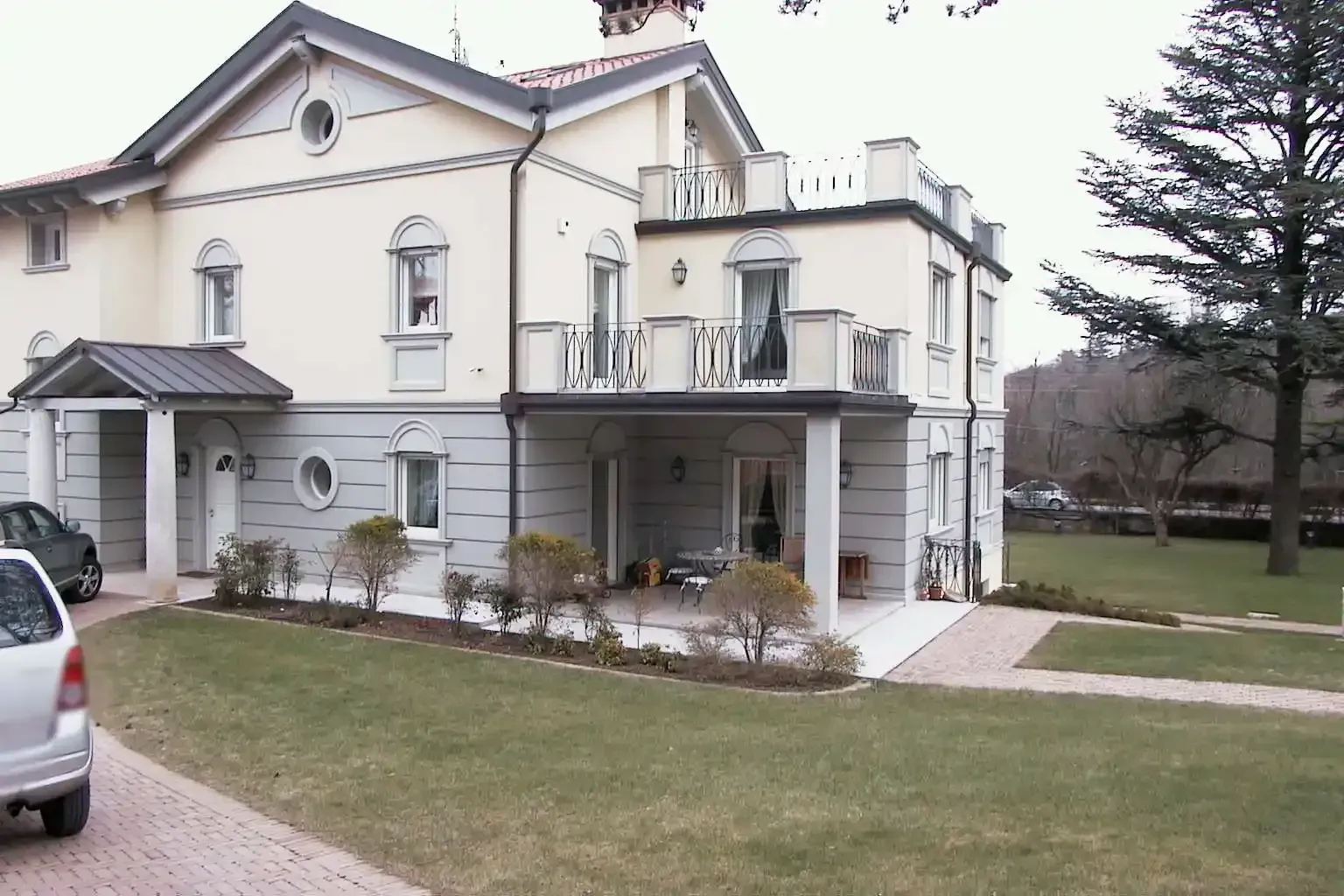 Una villetta suburbana a due piani, color crema e grigio, con balcone, veranda e un'auto parcheggiata sul vialetto di ghiaia.