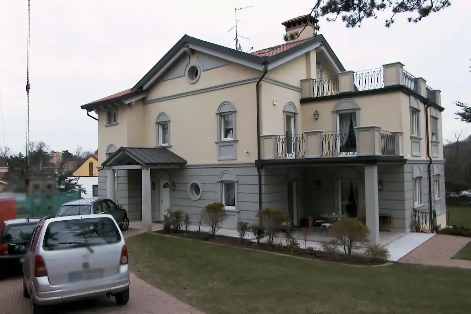 Una casa a due piani color beige e grigio con balcone, veranda e due auto parcheggiate su un prato.