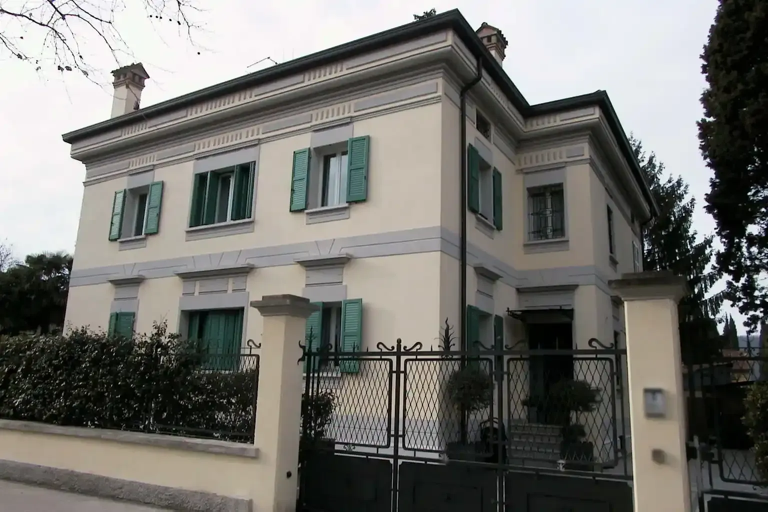 Una villa a due piani color crema con persiane verde chiaro, modanature decorative e un cancello in ferro battuto nero.