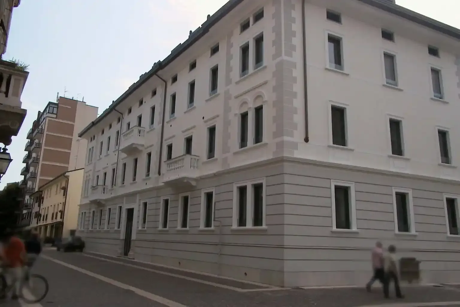 Un edificio bianco e grigio di tre piani si erge all'angolo di una strada, con pedoni e un ciclista che passano di lì.