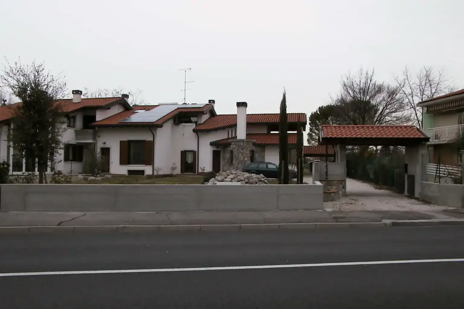 Una casa bianca a due piani con tetto in terracotta, camino in pietra e posto auto coperto laterale, vista da una strada adiacente.