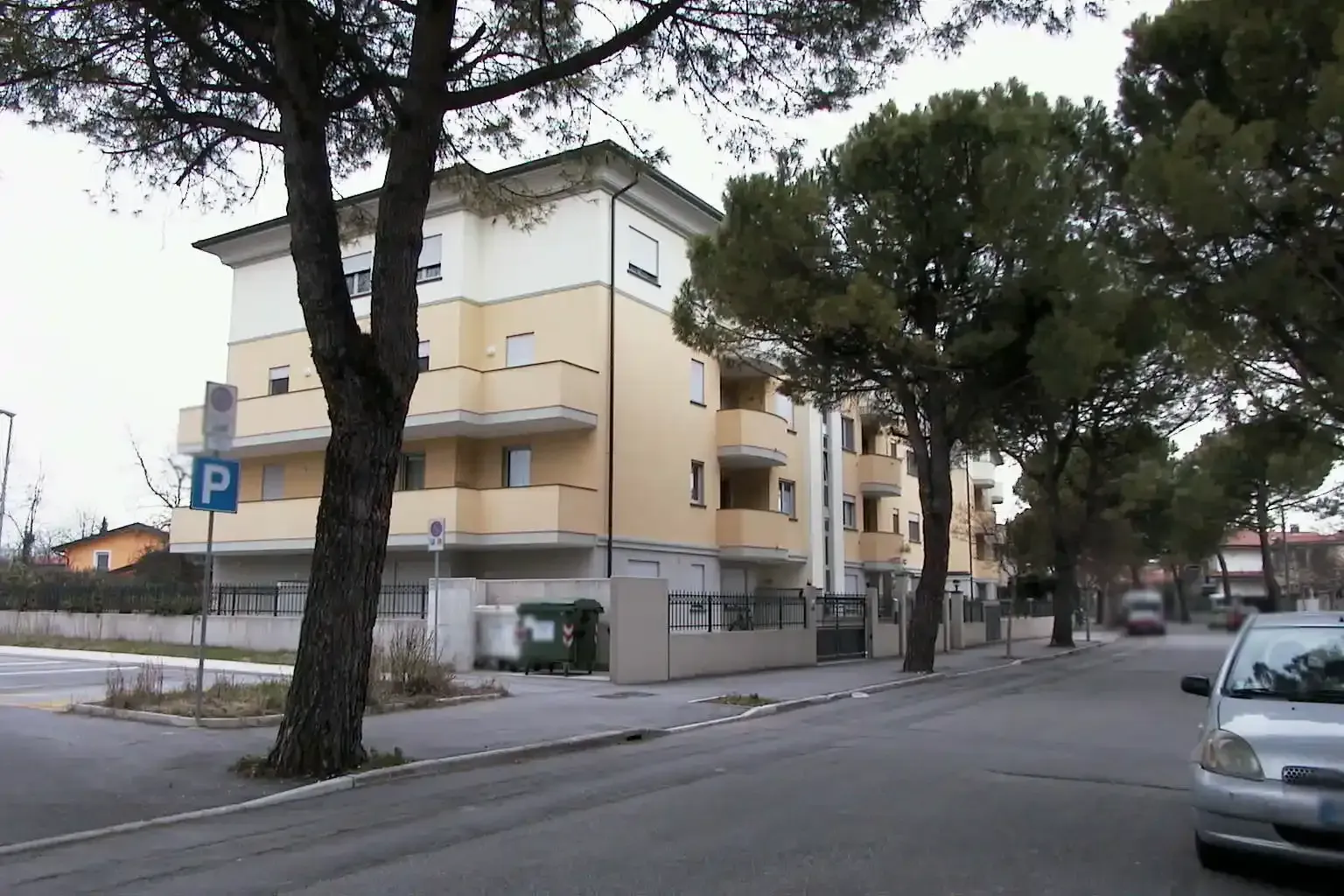 Un palazzo di tre piani di colore giallo pallido, visto dalla strada, fiancheggiato da pini in una giornata limpida e nuvolosa.