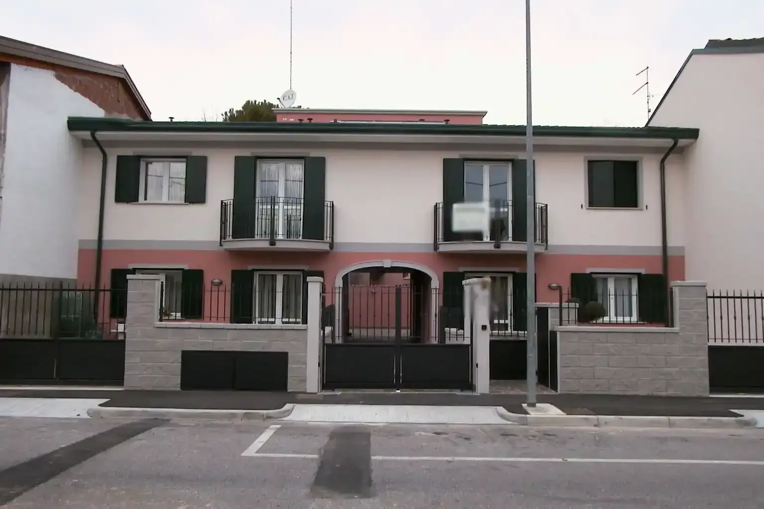Un edificio residenziale a due piani con il piano inferiore rosa, il piano superiore bianco, persiane verdi e un ingresso con cancello centrale.