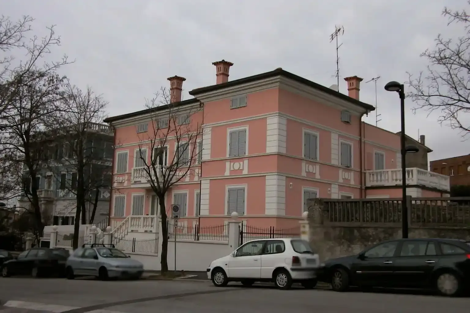 Una villa rosa a tre piani con cantonali bianchi si erge su una strada con auto parcheggiate sotto un cielo grigio e nuvoloso.