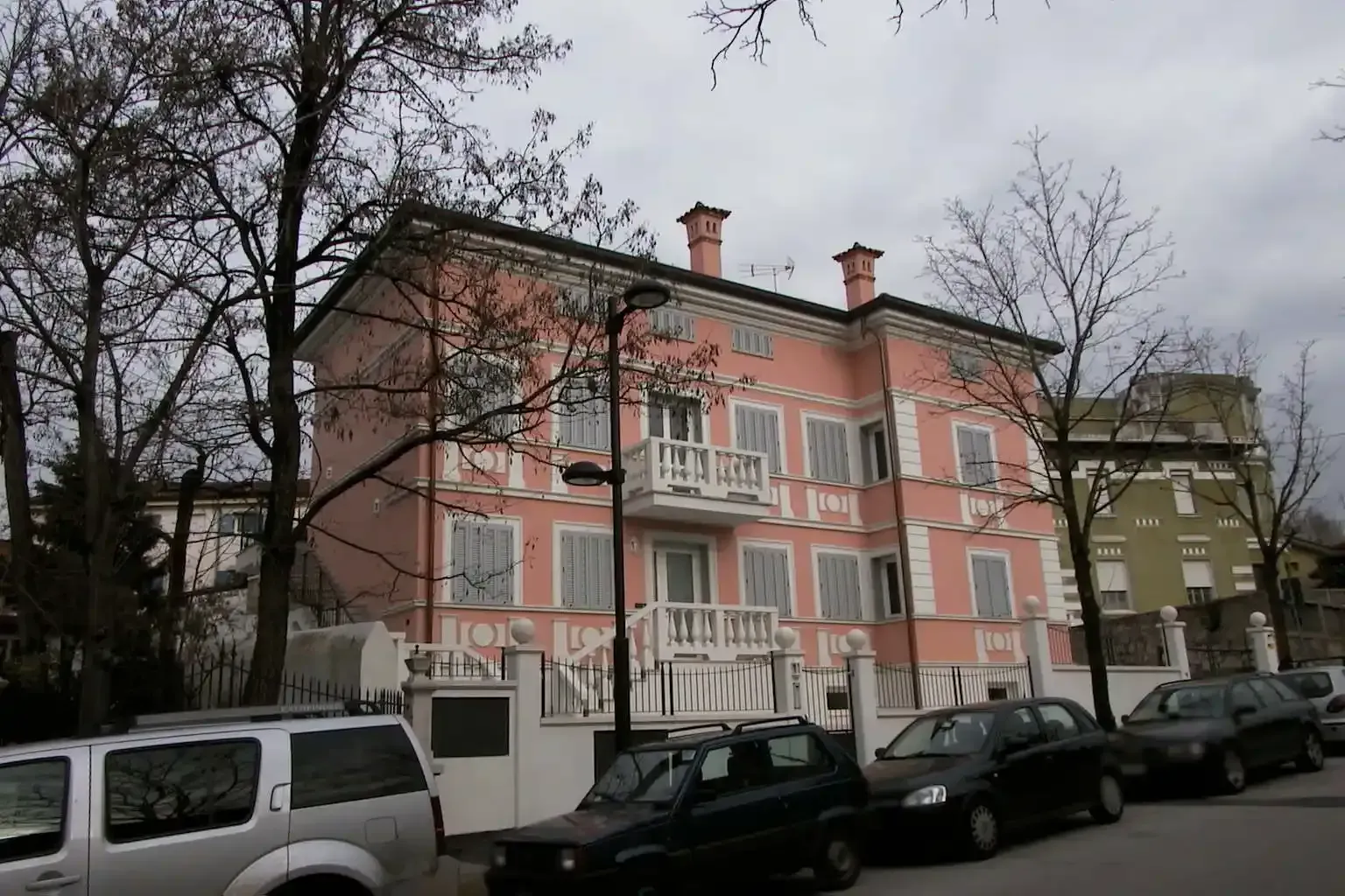 Un condominio rosa a tre piani con finiture e balconi bianchi, situato su una strada fiancheggiata da auto parcheggiate e alberi.