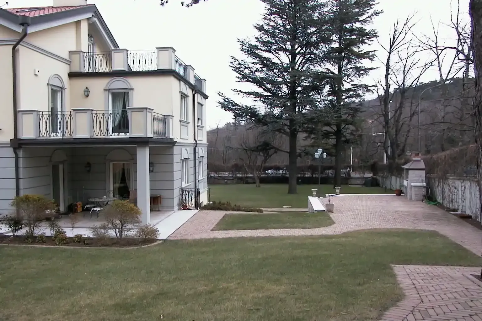 Una villa a due piani con balcone e patio, adiacente a un prato ben curato, vialetti lastricati e alti alberi sempreverdi.