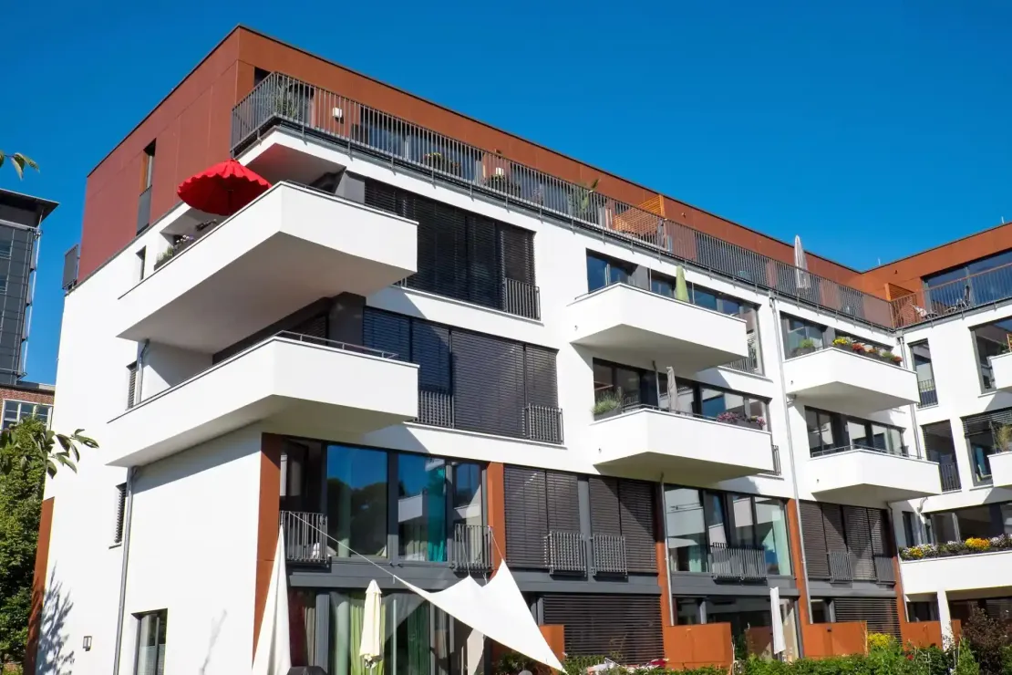 Un moderno condominio a più piani con balconi bianchi, ampie finestre e pannelli esterni marroni, sotto un cielo azzurro.