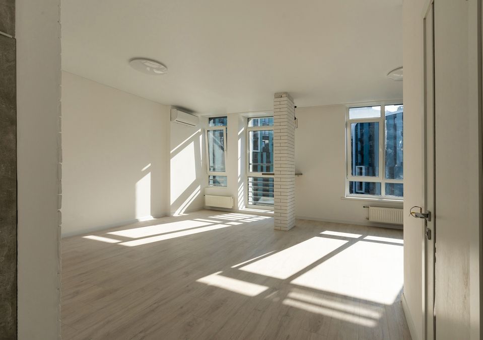 Una stanza vuota e soleggiata con pavimento in legno chiaro, pareti bianche, ampie finestre e un pilastro centrale in mattoni bianchi.