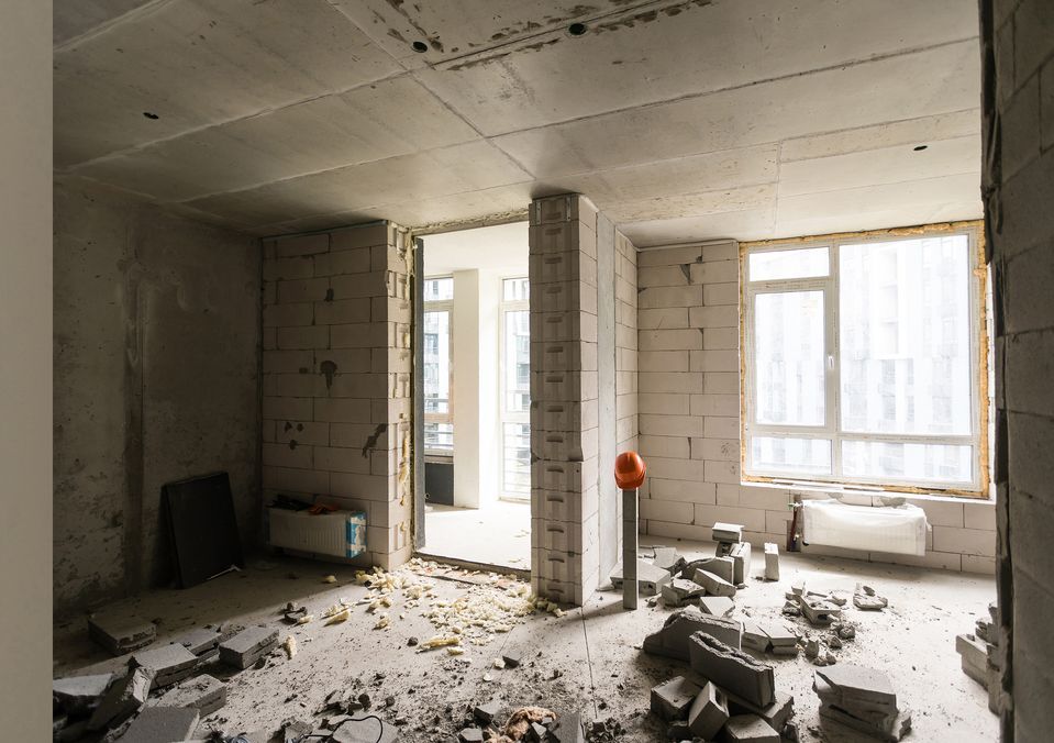 Una stanza non finita con pareti in blocchi di cemento a vista, detriti sul pavimento e una grande finestra.