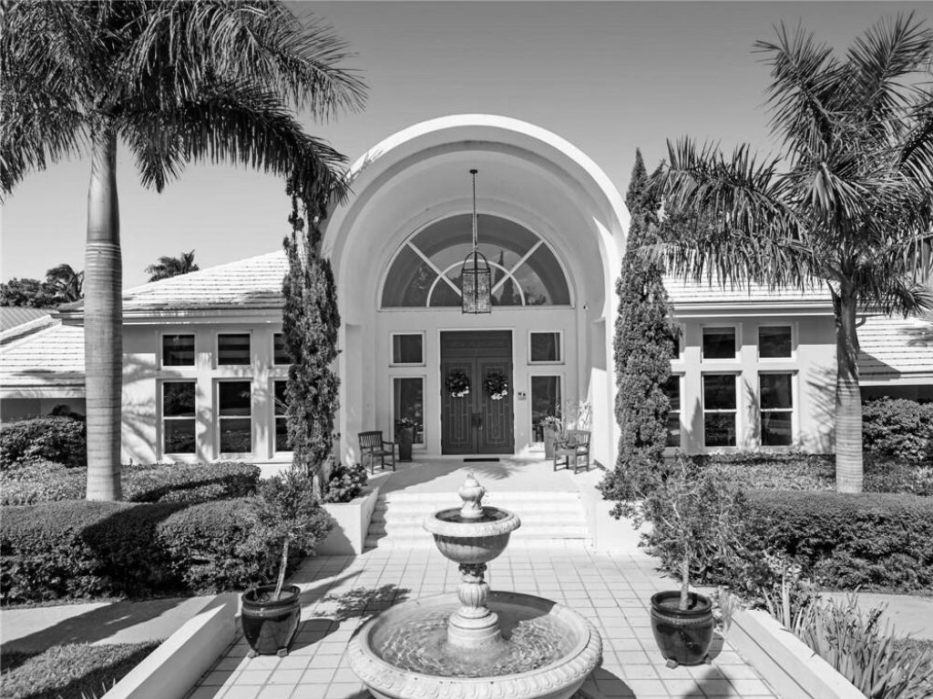 Grand arched entrance to a light-colored building, flanked by palm trees and shrubbery, with a fountain in the foreground.