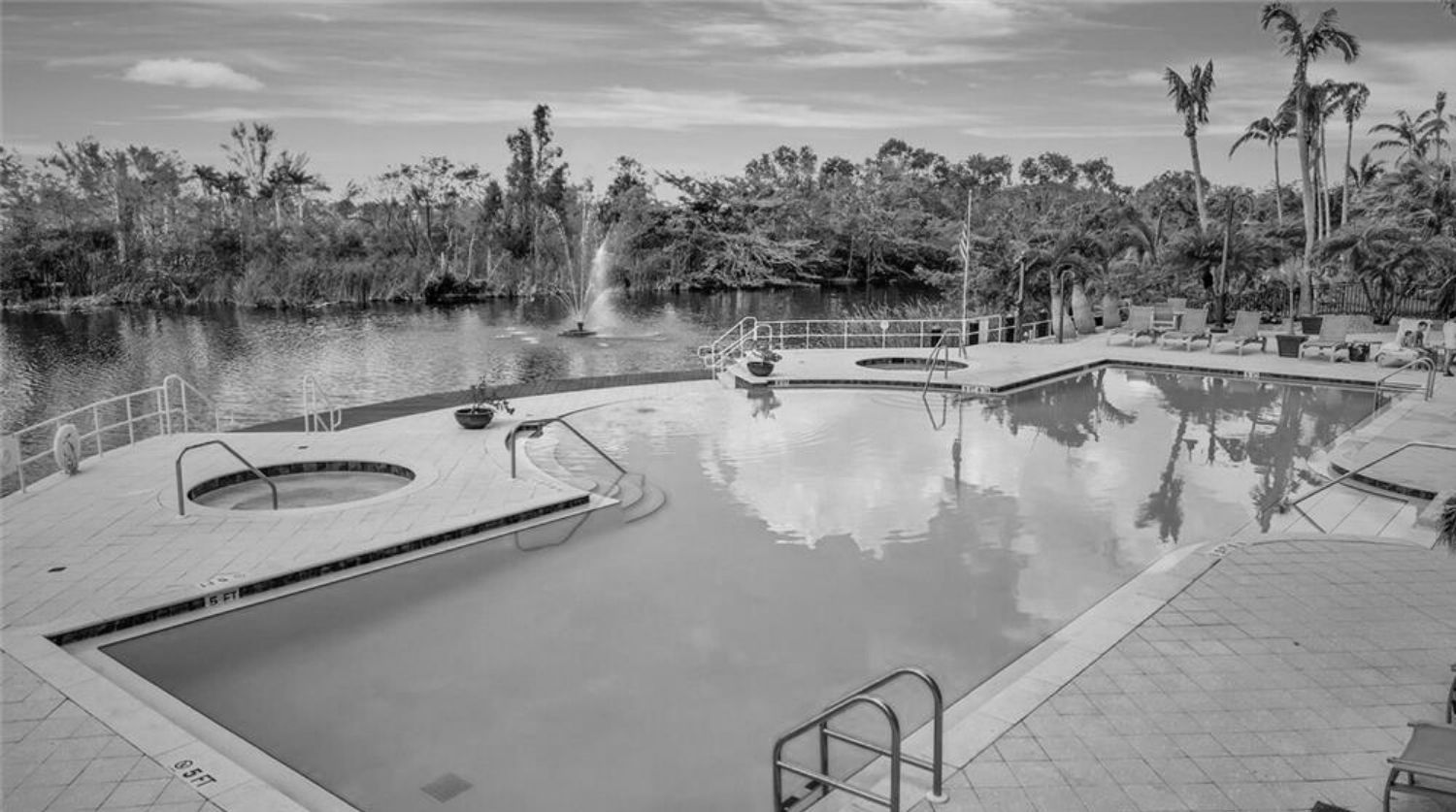 Black and white photo of a pool with a lake in the background, a fountain, and trees.