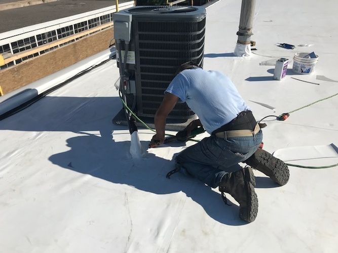 A man is kneeling on a white roof working on an air conditioner.