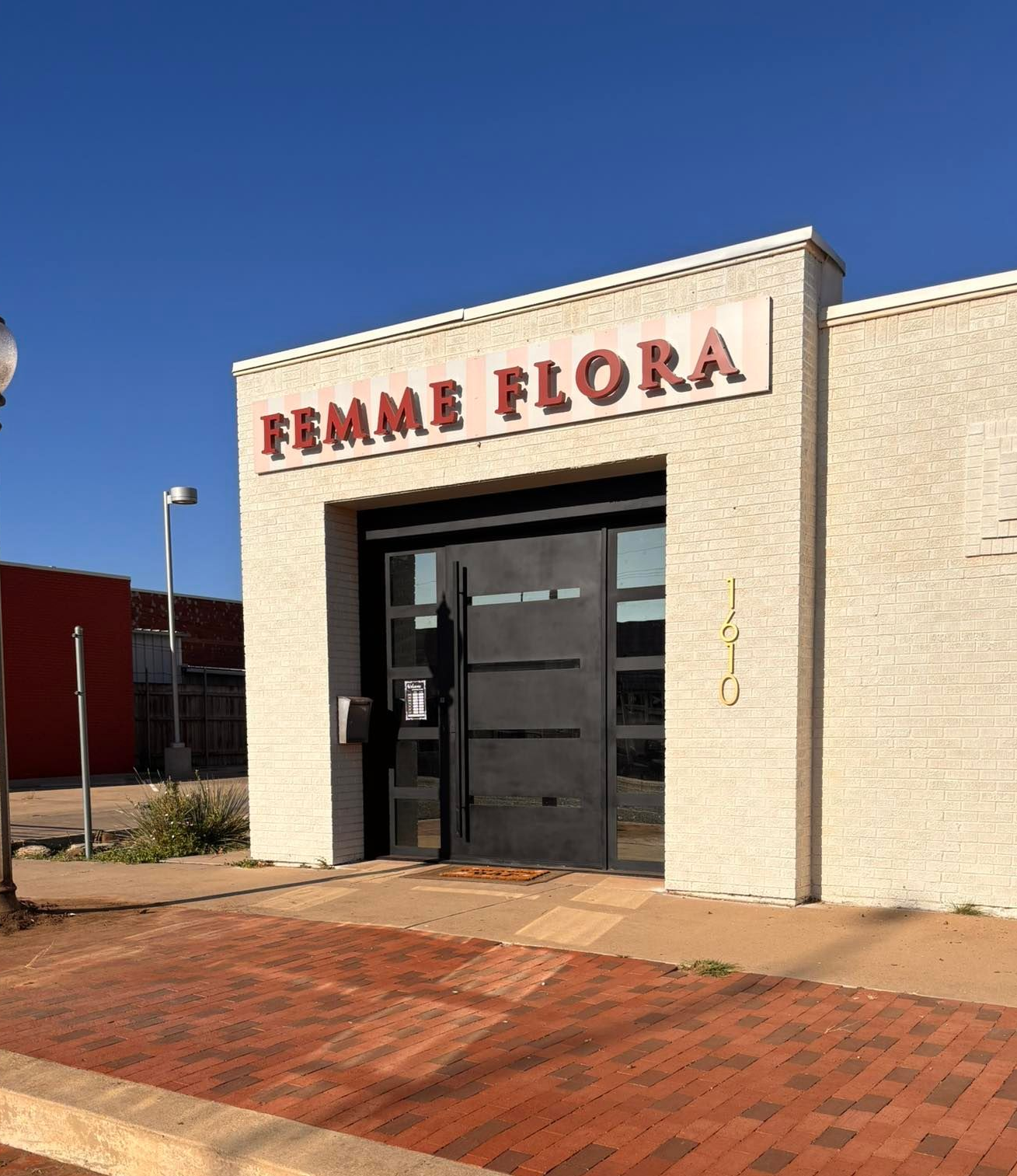 Femme Flora floral shop with black door, red brick sidewalk, and cream-colored building with red sign on a sunny day.
