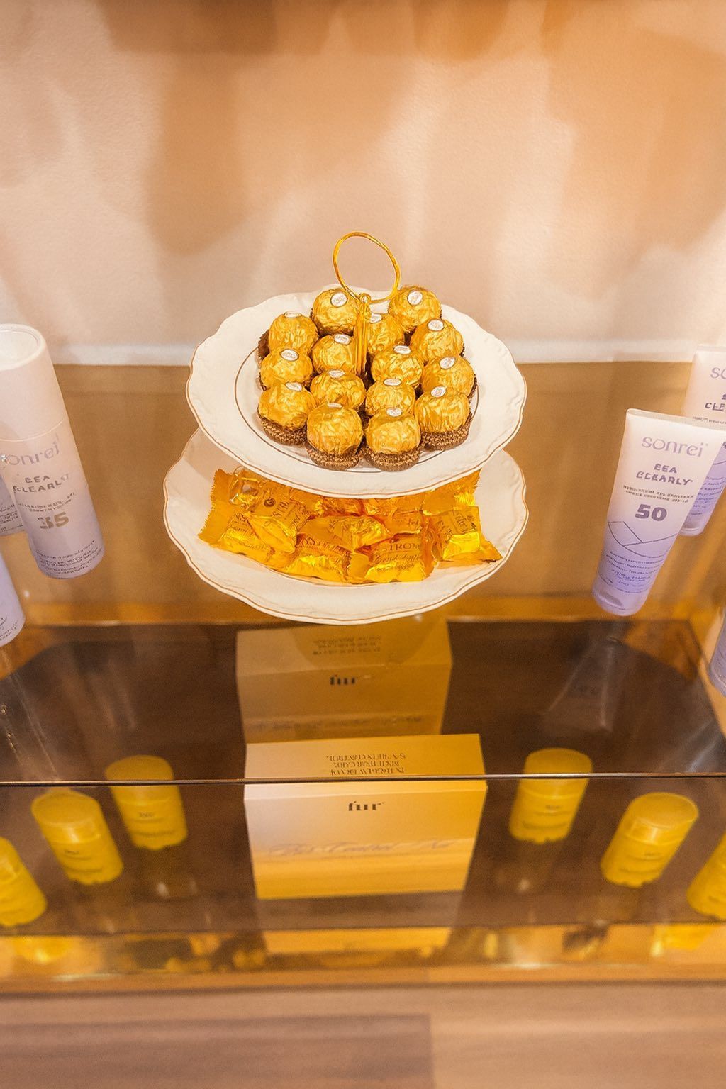 Display case with two-tiered white serving dish of golden treats. Beauty products surround, yellow and lavender.