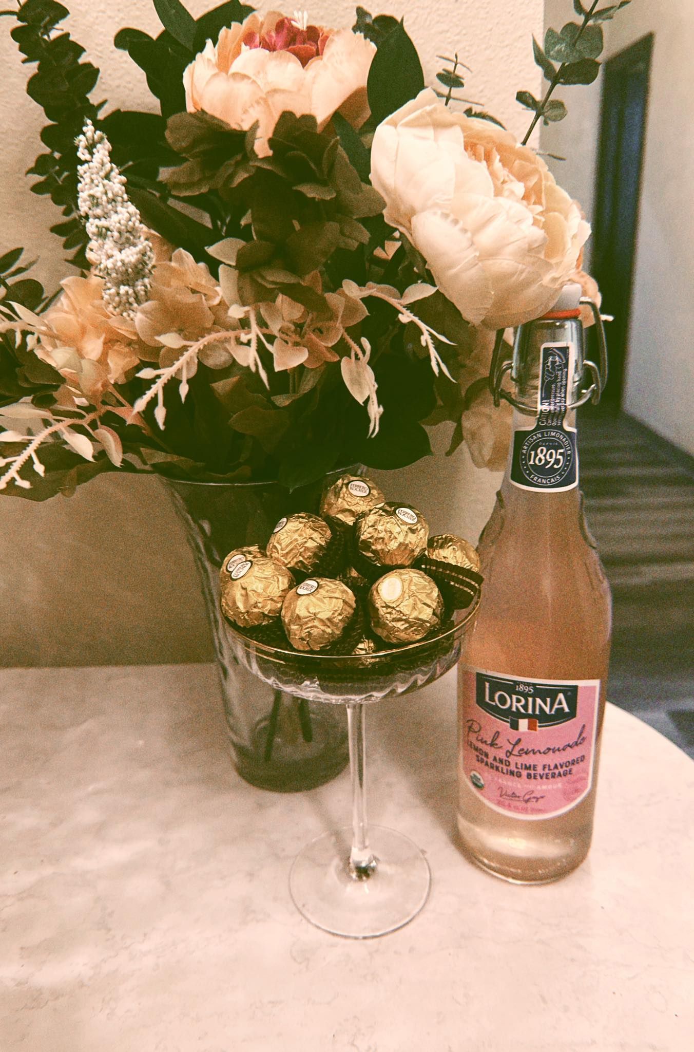 Flowers, chocolates, and a bottle of pink soda on a white table; soft lighting.