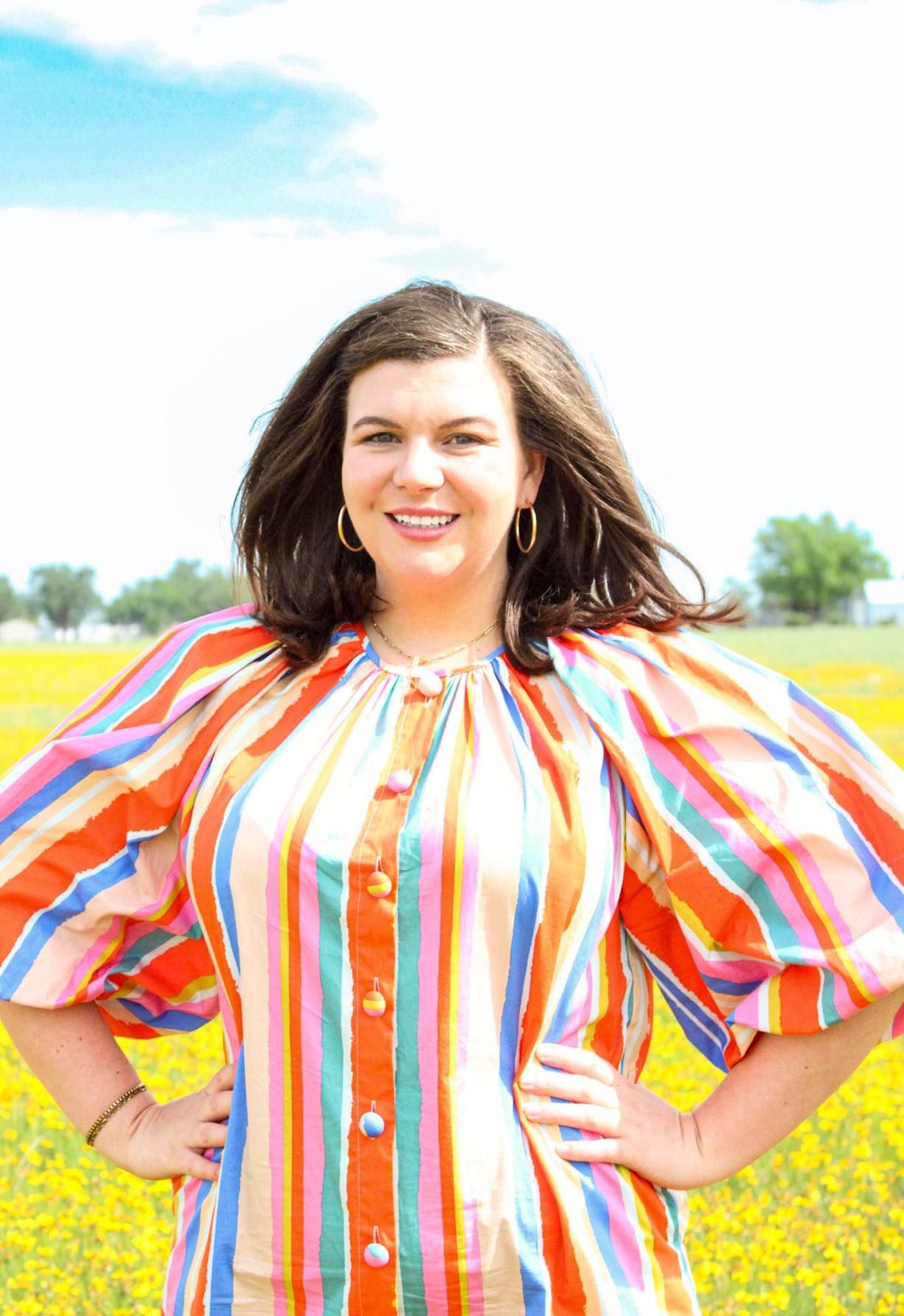 Woman in striped dress stands in field, arms on hips.