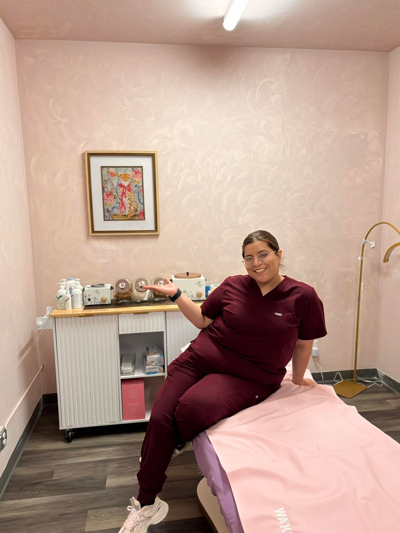 Woman in burgundy scrubs sits on a pink-covered spa bed, pointing, in a pink-walled room.