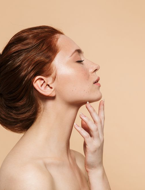Woman with red hair and freckles, touching her jawline, eyes closed, against a beige background.