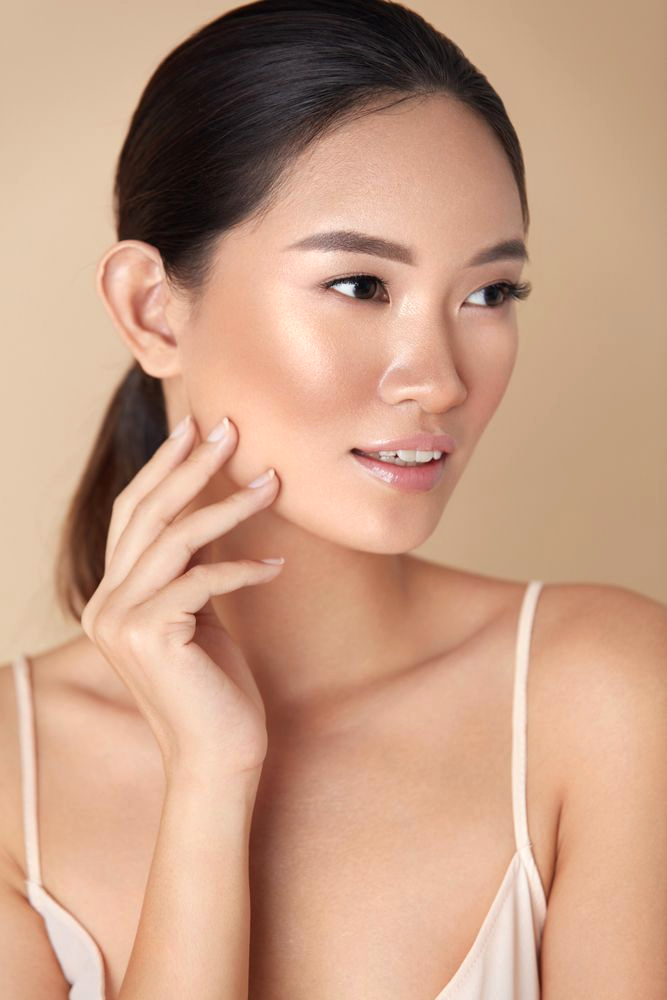 Woman with highlighted skin, touching her cheek, looking away; neutral background.