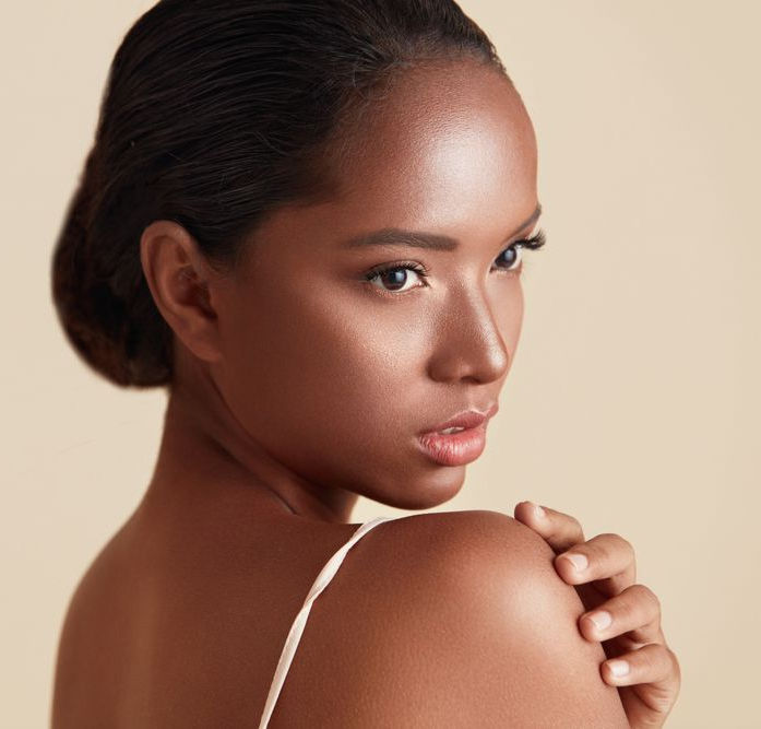 Woman with dark skin, looking over her shoulder, wearing a white strap top. Soft lighting, neutral background.