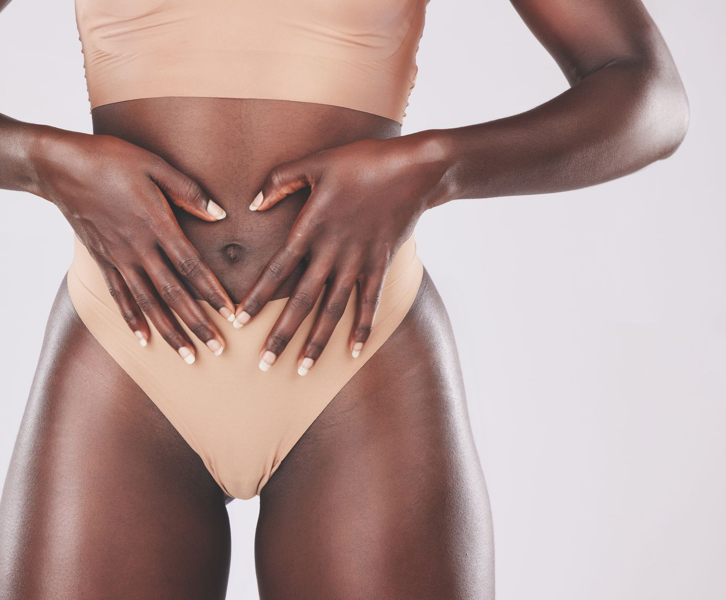 Woman making a heart shape on her stomach with her hands; wearing nude-colored underwear and top.