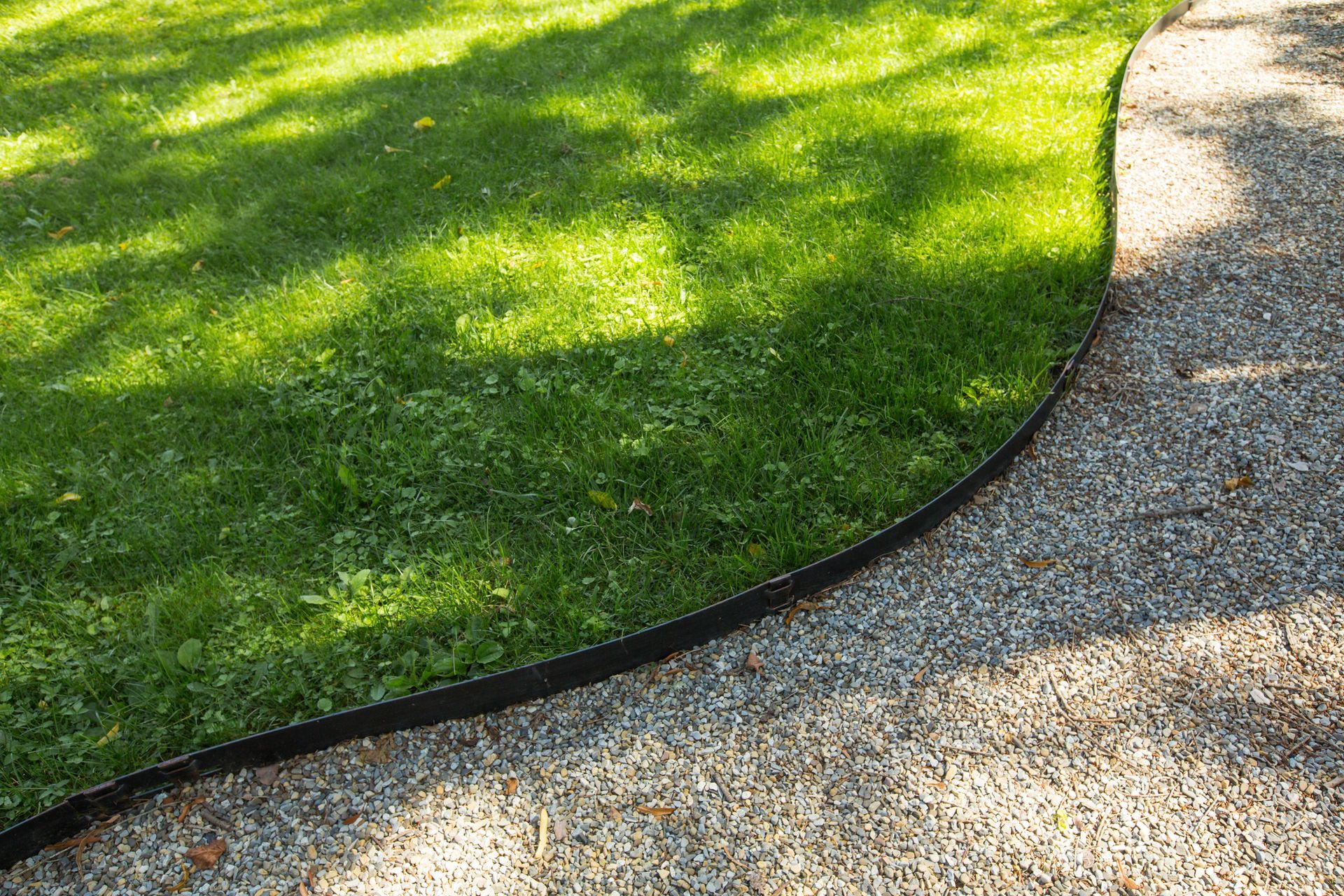 Lush green grass borders a gravel path, separated by a thin black edge. Sunlight casts shadows.