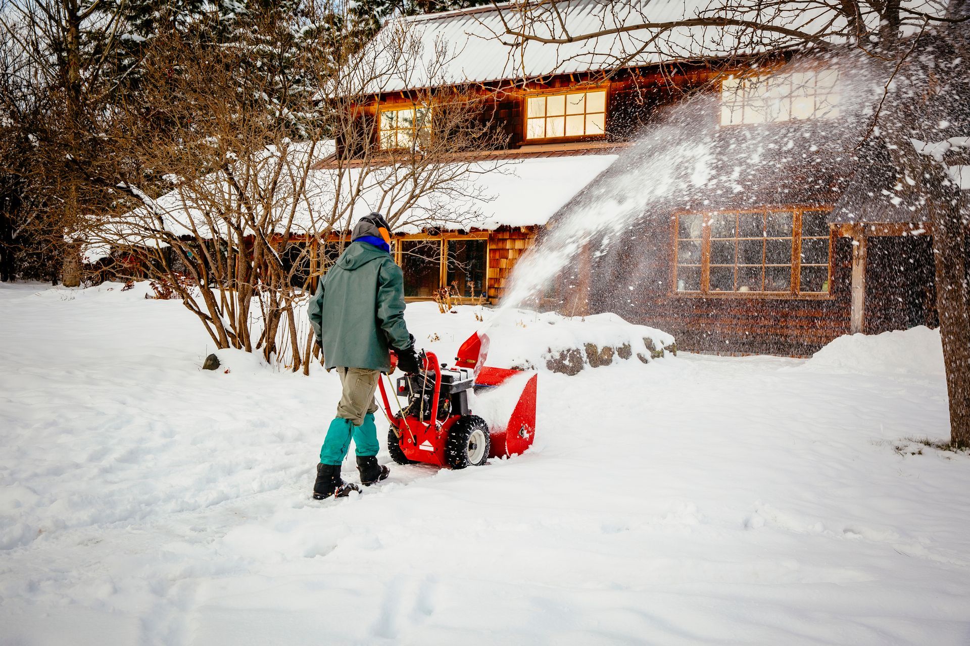 Person using a red snowblower clears snow near a brown house, winter setting.