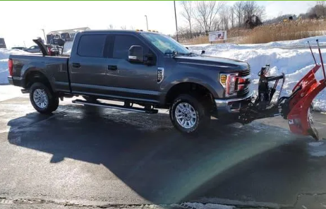 Dark gray pickup truck with snow plow in a snowy parking lot.