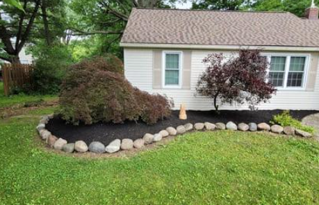 Small beige house with a curved rock border around a dark mulch flowerbed and two small trees.