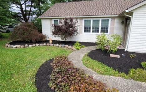 A house with white siding and brown roof. A curving walkway leads to the front door. Landscaping includes mulch beds, grass, and plants.