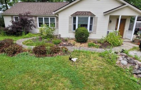 A beige house with a curved stone path leading to the front door, surrounded by a garden and overgrown grass.