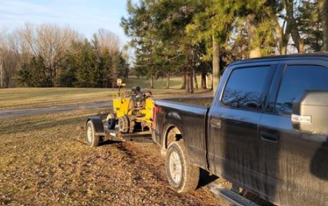 Black pickup truck towing a yellow machine on a trailer across a grassy field.