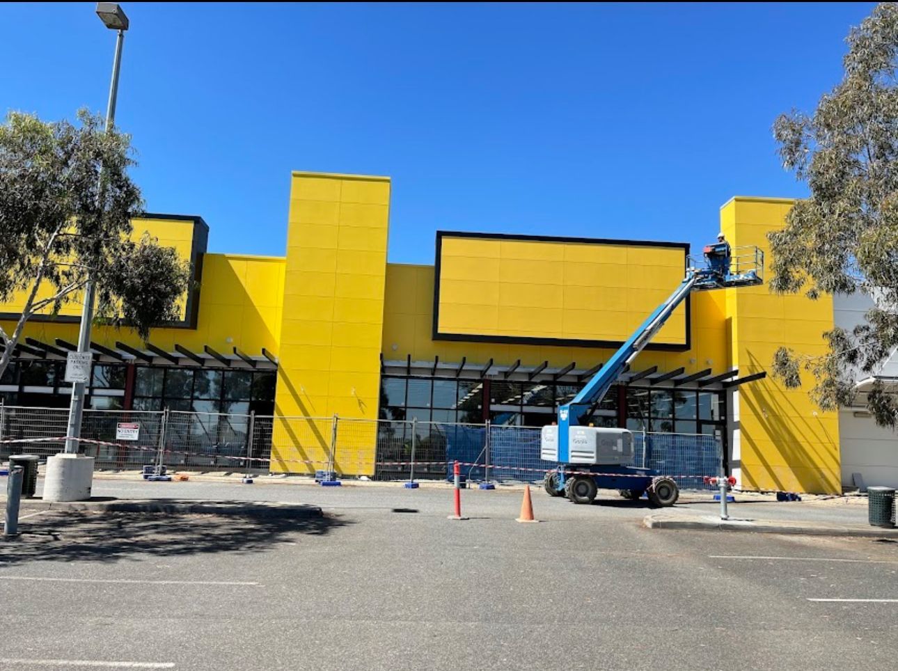 Yellow building undergoing renovations with a blue lift in front. Sunny day, visible construction materials, and safety barriers.