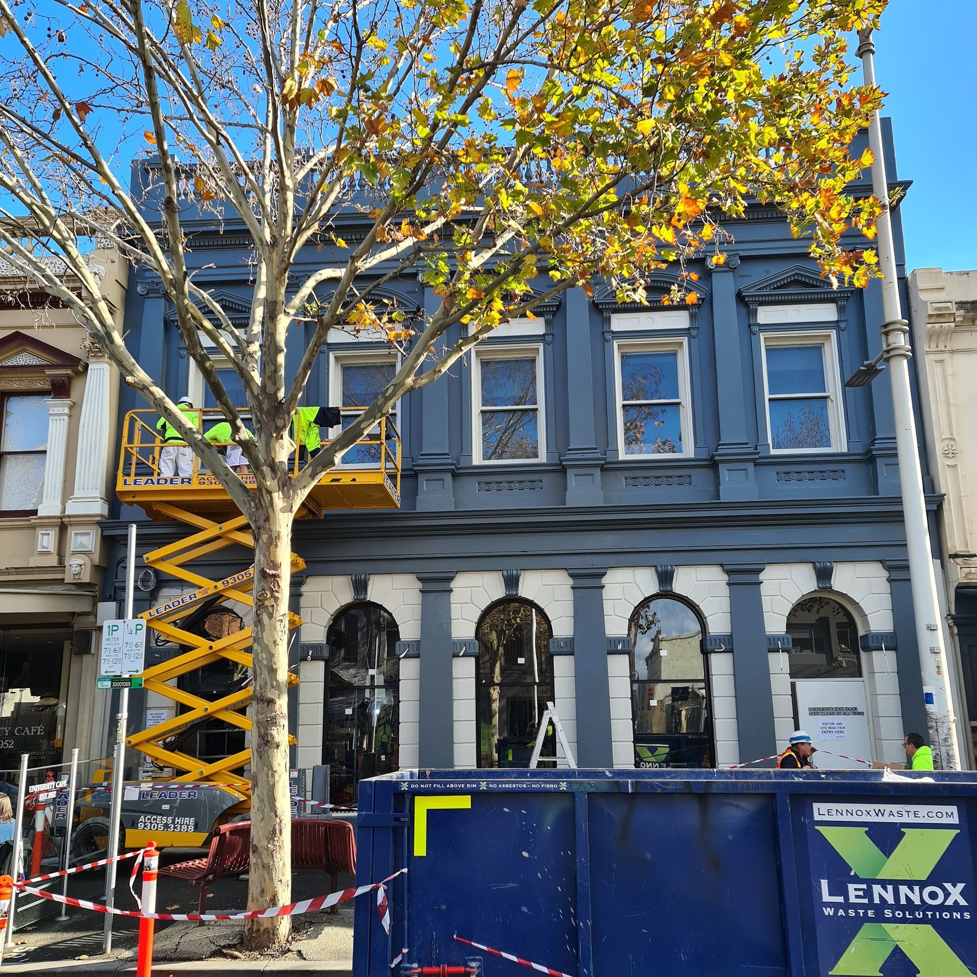 Historic blue building under renovation with a scissor lift and a dumpster in front, and a tree.
