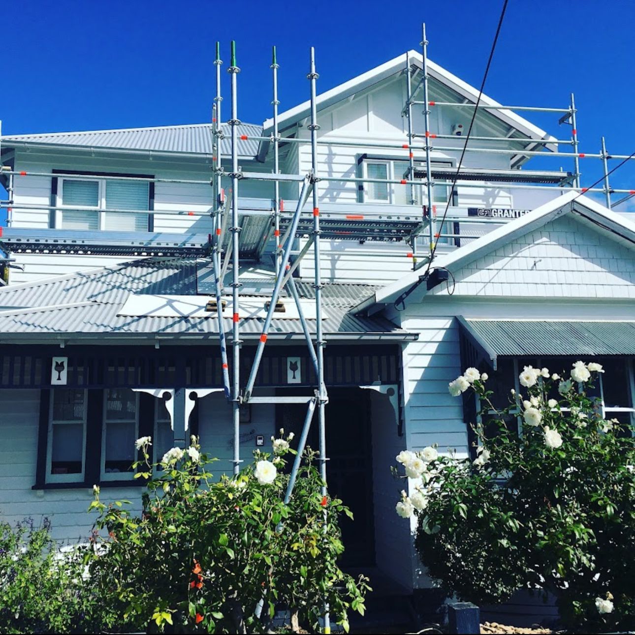 A white house with scaffolding on its roof, under a bright blue sky. Roses bloom in the foreground.
