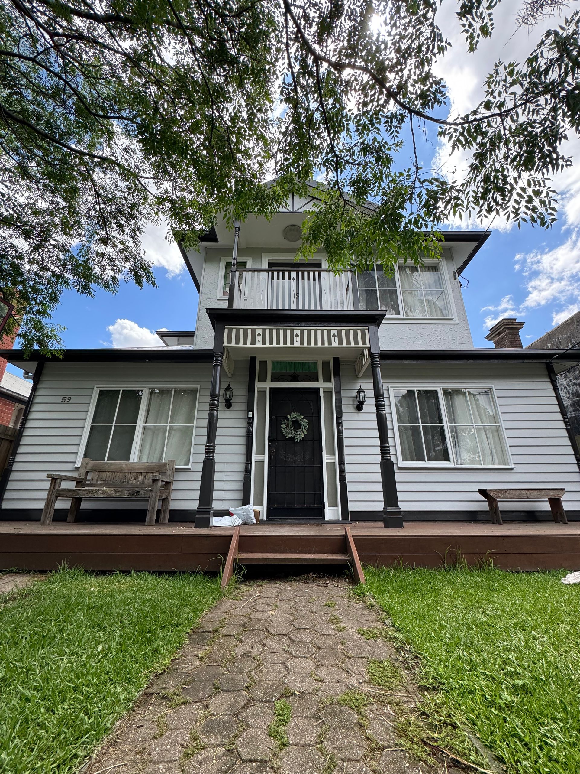 A two-story house with white siding and a black door and trim. A wooden porch and walkway lead to the front door under trees.