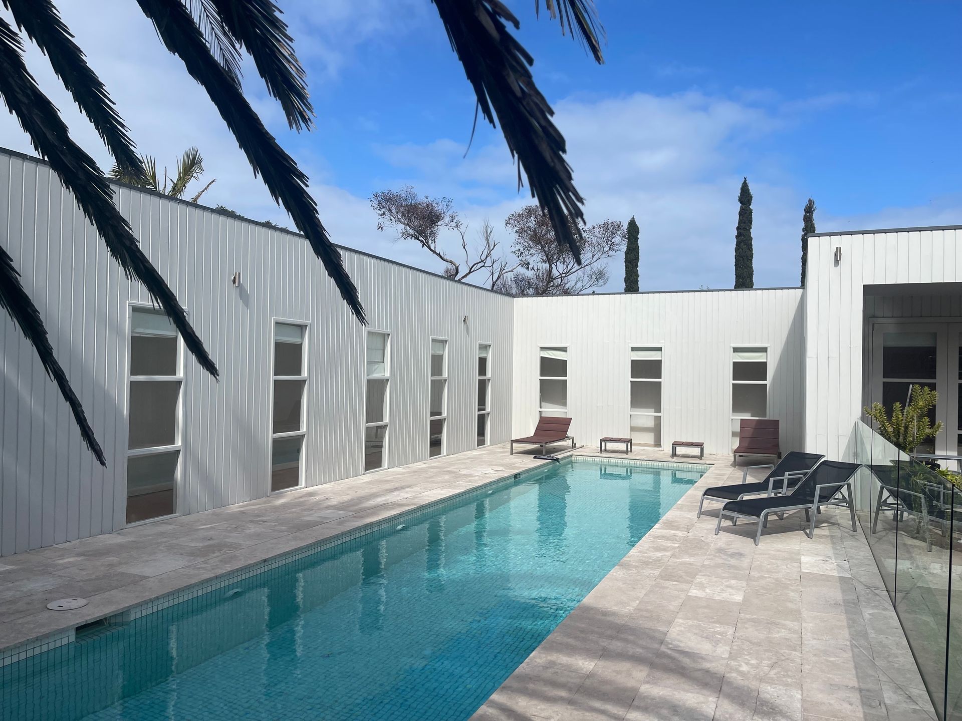 Pool area with long, rectangular pool bordered by white buildings and lounge chairs, under a bright blue sky.