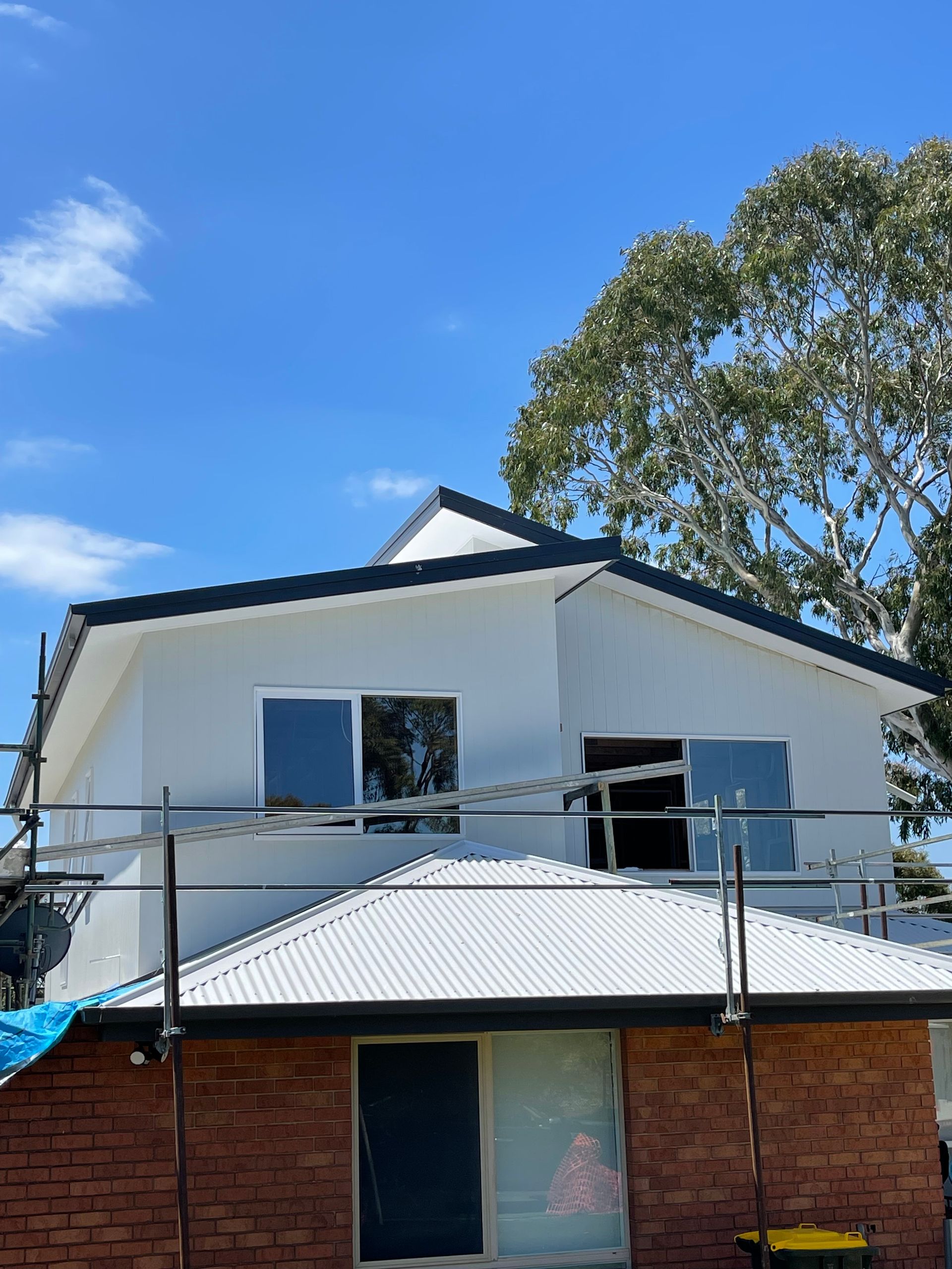 Two-story house under construction. White upper level with black roof trim, brick lower level. Scaffolding and a tree present.