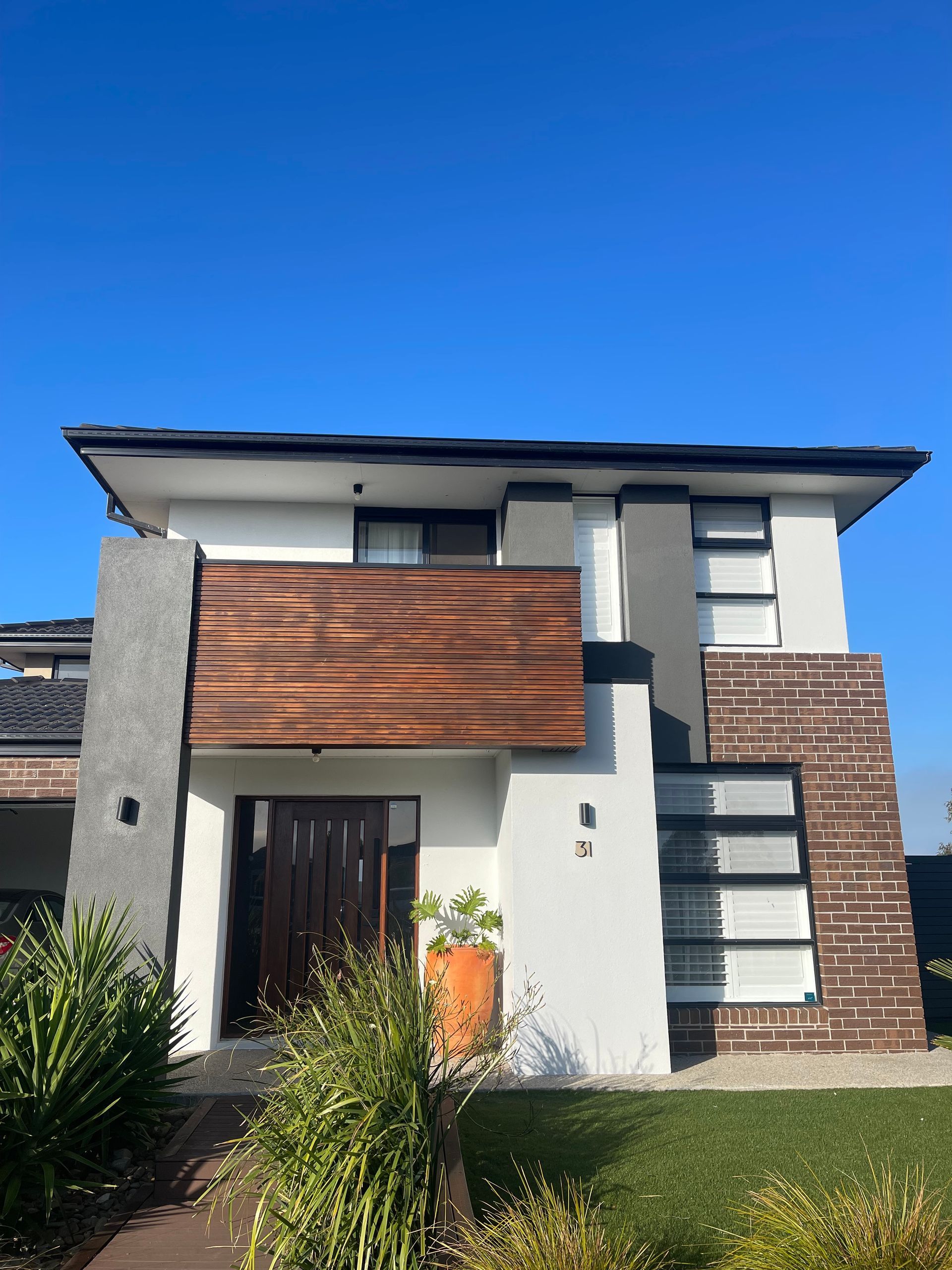 Two-story modern home with white walls, a wooden balcony, and brick accents against a blue sky.