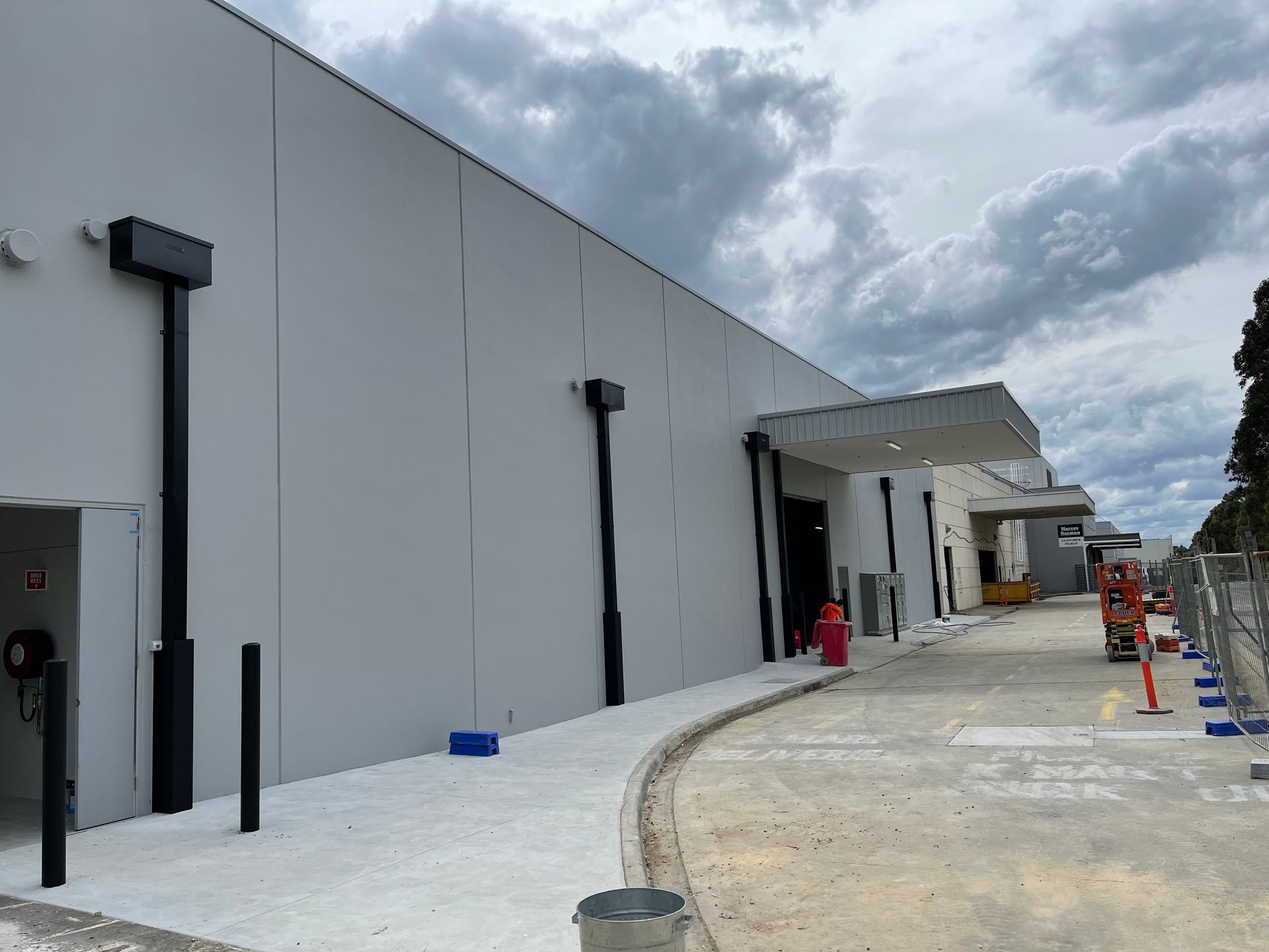 Exterior of a light gray industrial building under a cloudy sky. A sidewalk and construction barriers are in the foreground.