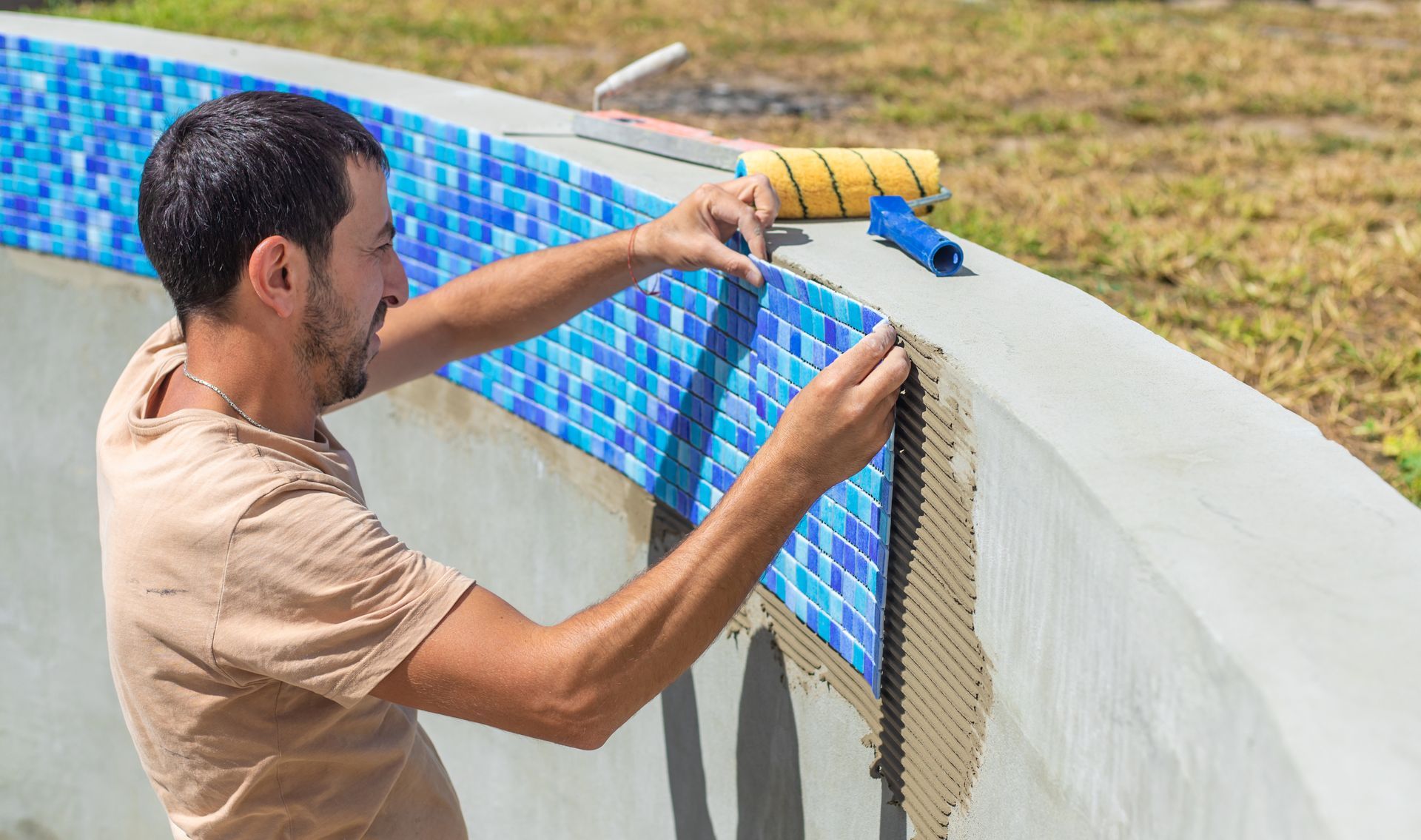 Man tiling the edge of a pool with blue mosaic tiles.