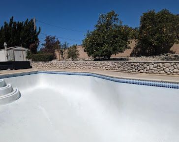 Empty white swimming pool with blue tile trim, rock wall, and trees against a blue sky.
