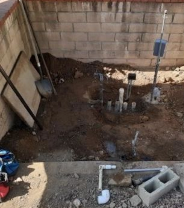 Construction site: exposed pipes and dirt in a walled area, tools leaned against wall.