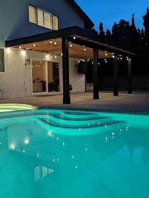 Poolside patio with string lights, illuminated pool, and house at dusk.
