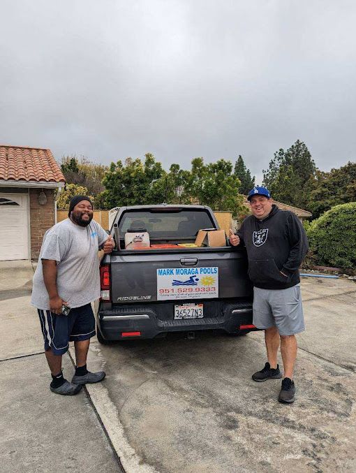 Two men stand beside a truck with food boxes, a house in the background, cloudy sky.