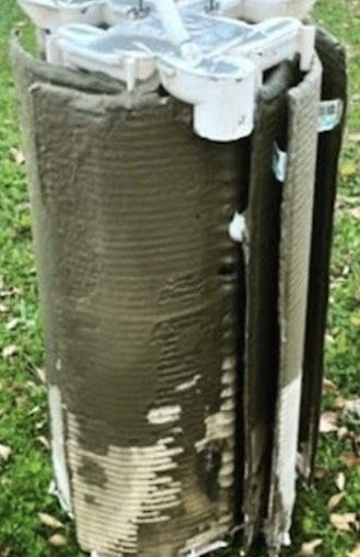 Clogged water filter; green-brown residue covers the cylindrical structure. Outdoor setting, grassy background.