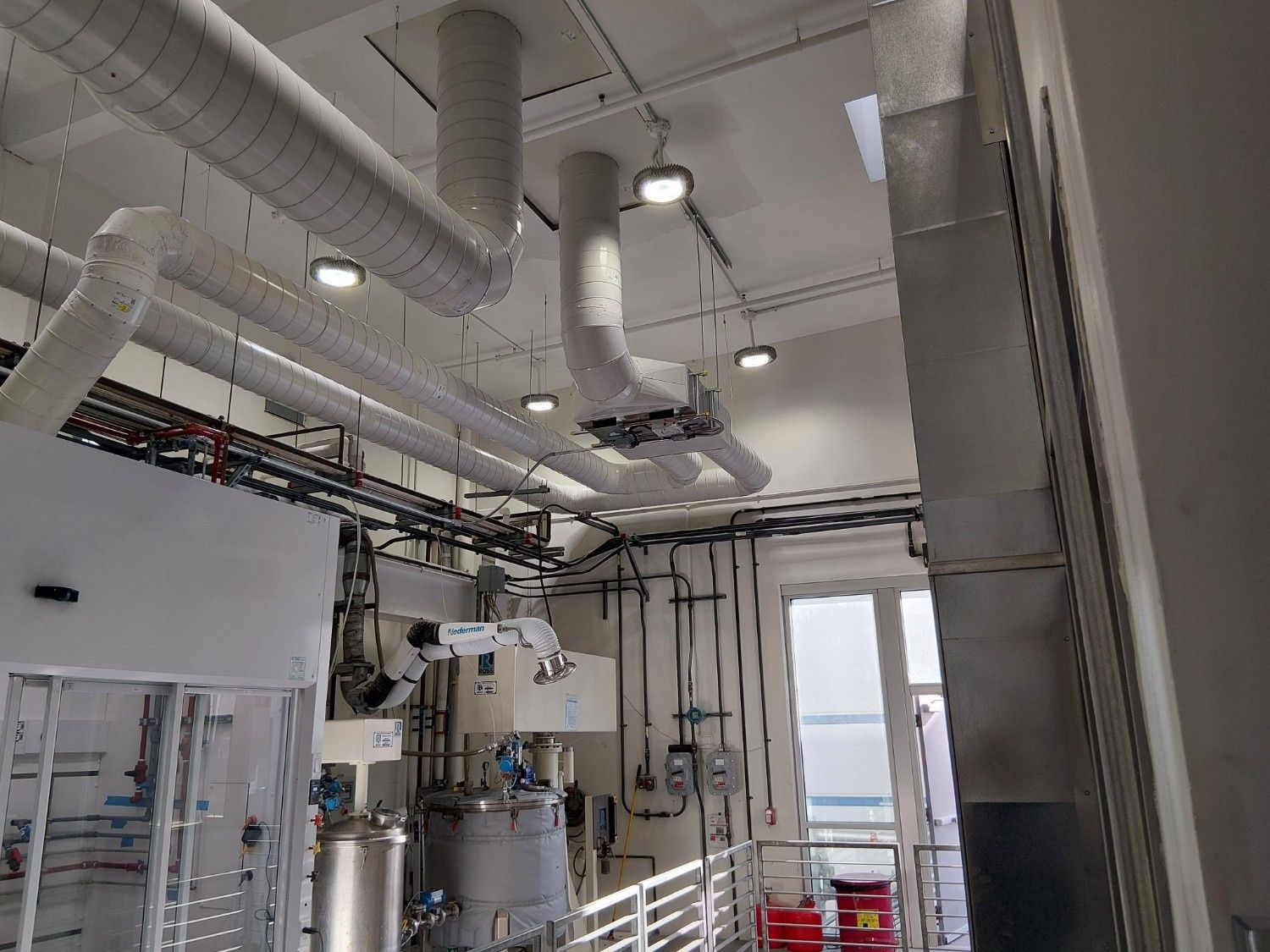 A laboratory interior featuring industrial machinery, silver stainless steel equipment, and overhead ventilation ducts.