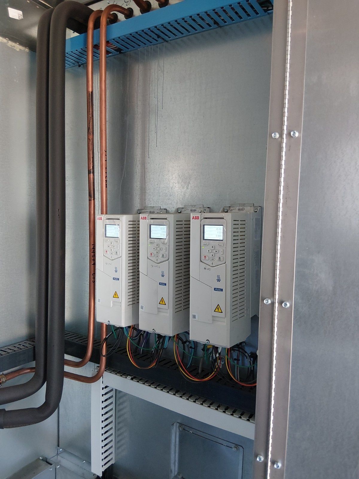 Three white variable frequency drives mounted on a gray wall inside a metal enclosure with copper piping.