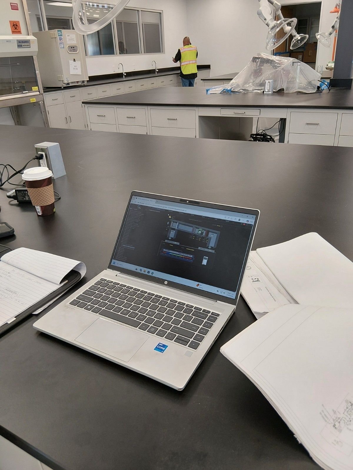 A silver laptop on a black lab bench with notebooks, a coffee cup, and a person in a yellow vest in the background.