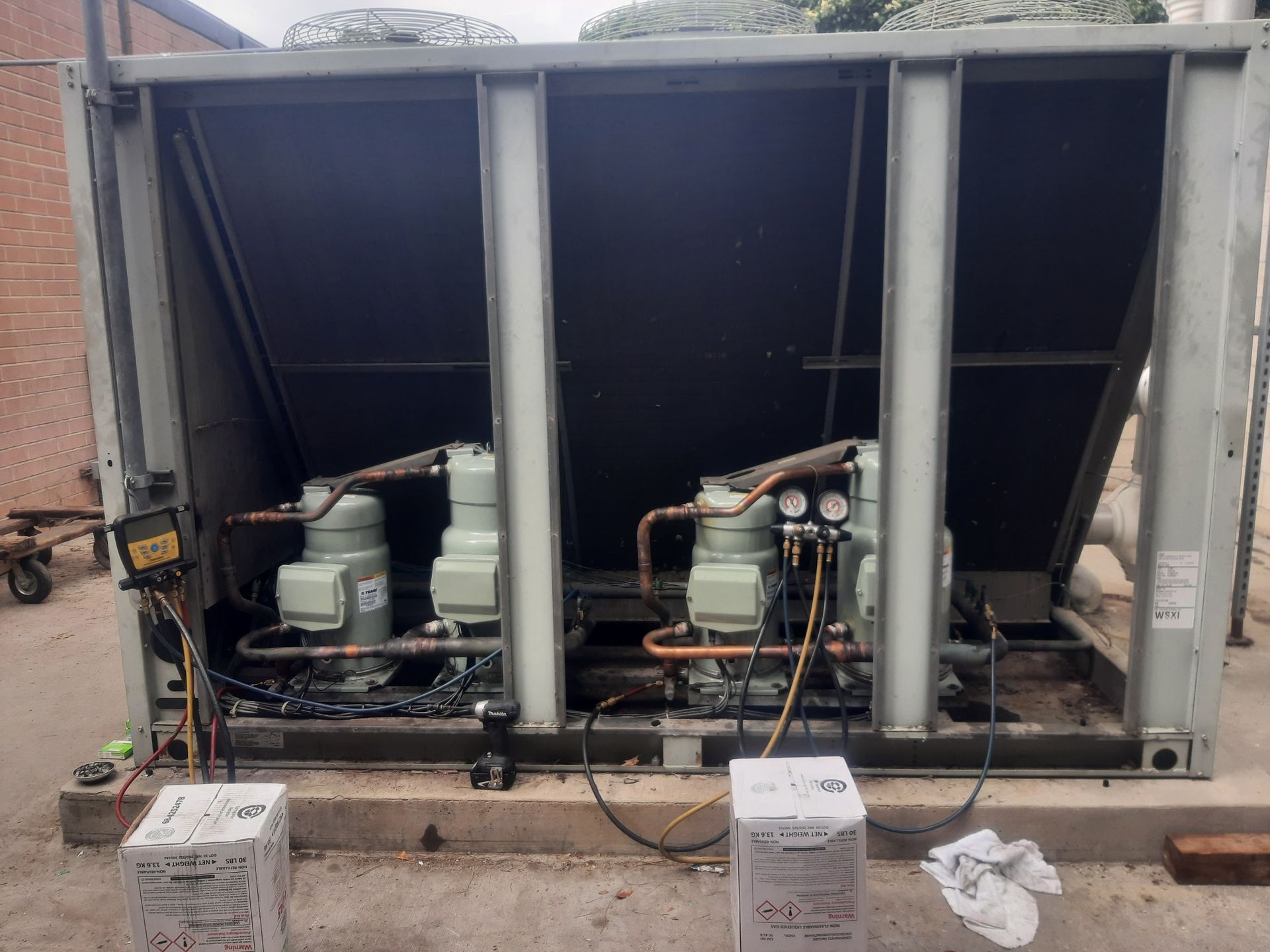 An outdoor commercial air conditioning unit with two visible compressors undergoing service, with gauges attached.